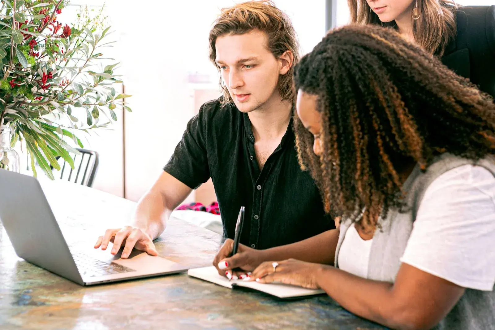 3 people working together at a table, one using a laptop, one writing in a notebook, the third standing behind